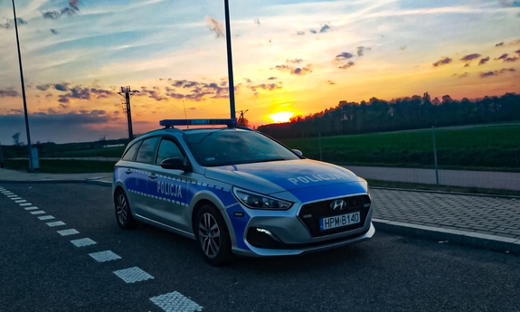 Radiowóz zaparkowany na poboczu drogi, w tle las i zachodzące słońce.