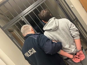 Policjant prowadzi zatrzymanego mężczyznę w kajdankach.