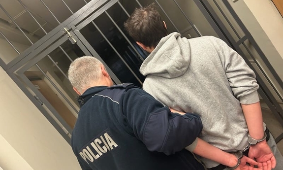 Policjant prowadzi zatrzymanego mężczyznę w kajdankach.