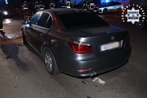 Tył pojazdu marki BMW w kolorze szary metalik, w tle pojazd Straży Pożarnej z włączonymi światłami błyskowymi w kolorze niebieskim.