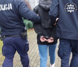 Dwaj policjanci prowadzą kobietę trzymając ją za ręce. Kobieta ma ręce trzymane z tyłu spięte kajdankami.