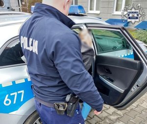 Policjant stoi przy prawych tylnych drzwiach radiowozu, do którego wsiada kobieta.