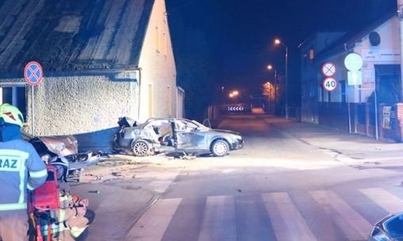 Miejsce wypadku w Praszce na skrzyżowaniu ulic Powstańców Ślaskich i Wodnej. Na pierwszym planie w prawym dolnym rogu stojący na jezdni pojazd policyjny z włączonymi światłami błyskowymi w kolorze niebieskim. W oddali po lewej stronie kadru dwa rozbite pojazdy. Po lewej i prawej stronie obecni na zdjęciu Strażacy - Ratownicy w mundurach w kolorze piaskowym.