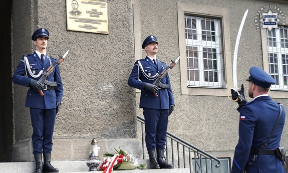 Policjant oddaje honor, w tle na baczność stoi dwóch policjantów.