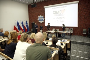 Na podeście w Sali konferencyjnej Dyrektor Gabinetu Komendanta Głównego Policji wita gości - dwie kobiety, przedstawicielki Fundacji Feminoteka. Obok stoi Pełnomocniczka Komendanta Głównego Policji ds. wdrażania Planu Równości Płci w Policji na lata 2023-2026. Na pierwszym planie uczestnicy spotkania. Za osobami na podeście wyświetla się na rzutniku napis Witamy na szkoleniu. Stand Up. Sprzeciw się molestowaniu w miejscach publicznych, od L’Oréal Paris, Right To Be, Feminoteka.