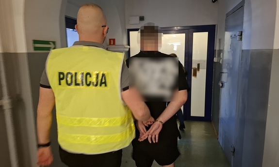 Policjant prowadzi zatrzymanego mężczyznę zakutego w kajdanki.