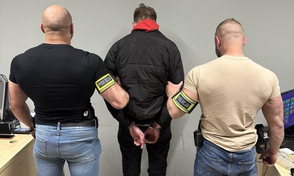 Dwaj nieumundurowani policjanci oraz zatrzymany mężczyzna z kajdankami założonymi na ręce z tyłu. Widok z tyłu.