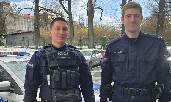 dwóch policjantów pozuje do zdjęcia. w tle stoi radiowóz.