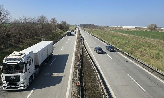 Zdjęcie przedstawia drogę czteropasmową, po której jadą ciągniki siodłowe i pojazdy osobowe. Po obu stronach drogi znajdują się pola i drzewa.