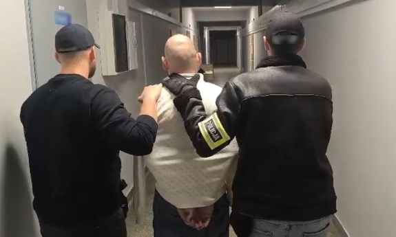 Na zdjęciu dwóch nieumundurowanych policjantów, z których jeden ma założoną na ręku żółtą opaskę z napisem POLICJA, i pośrodku nich zatrzymany mężczyzna. Policjanci prowadzą go korytarzem.