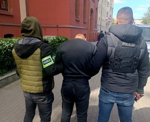 Dwaj funkcjonariusze prowadzą zatrzymanego męczyznę.