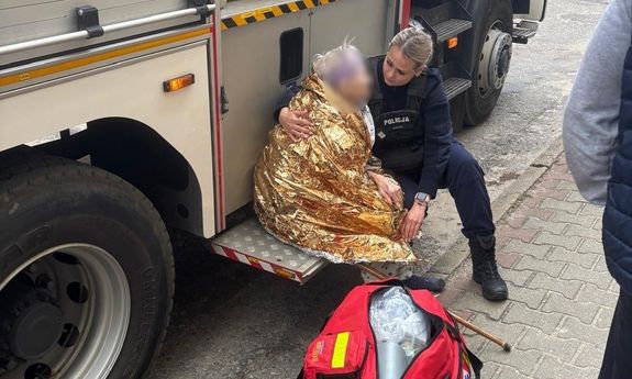 Policjantka przytula owiniętą kocem starszą panią siedzącą na platformie samochodu.