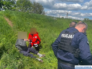 Ratownik medyczny podaje mężczyźnie siedzącemu na wzgórzu wodę obok policjanci.