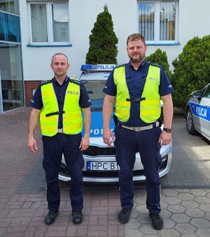 Dwaj policjanci stoją przed radiowozami zaparkowanymi przed budynkiem komendy.