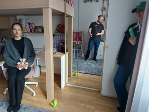 Młoda dziewczyna siedzi na krześle. Obok stoi kobieta, w głębi, w drugim pomieszczeniu mężczyzna.