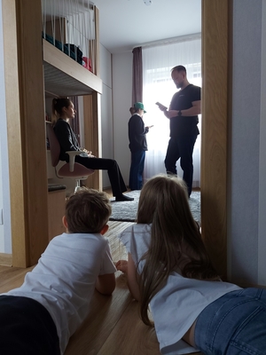 Dwójka dzieci leży na podłodze patrząc na osoby w drugim pomieszczeniu: młodą dziewczynę siedzącą na krześle, mężczyznę stojącego naprzeciwko niej, oraz dziewczynkę wyglądającą przez okno.