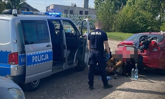 Policjant przy radiowozie. Obok na ziemi lezy zatrzymany mężczyzna, któremu policjant zakłada kajdanki.