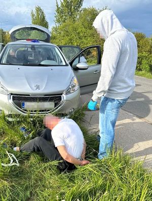 Funkcjonariusz z zatrzymanym siedzącym na skraju drogi, za nimi w głębi przy drodze pojazdy.