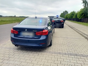 Na poboczu drogi stoi zaparkowany granatowy samochód, przed nim stoi auto z oznaczeniem na dachu literą L.