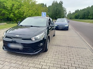 Na poboczu drogi stoi zaparkowane auto koloru czarnego z oznaczeniem na dachu literą L, za nim stoi granatowy samochód osobowy.