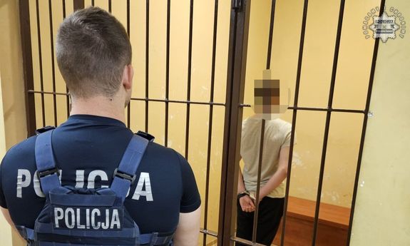 Policjant stoi przed kratami pomieszczenia w którym przebywa zatrzymany w kajdankach.