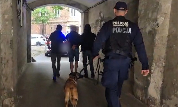 Dwaj policjanci prowadzą zatrzymanego mężczyznę, za nimi idzie policjant przewodnik z psem policyjnym.