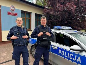 Policjant i policjantka stoją przy radiowozie policyjnym.
