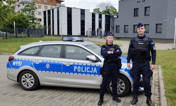 Dwoje umundurowanych policjantów przed radiowozem pozuje do zdjęcia.