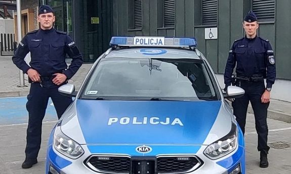 policjanci stoją obok radiowozu.
