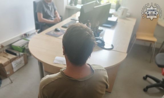 Zatrzymany mężczyzna siedzi na krześle - widok z tyłu. Przed nim widać siedzącego za biurkiem mężczyznę.