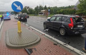 Miejsce kolizji drogowej. Uszkodzony samochód osobowy. W głębi widoczny radiowóz policyjny i inne samochody osobowe.