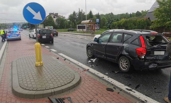 Miejsce kolizji drogowej. Uszkodzony samochód osobowy. W głębi widoczny radiowóz policyjny i inne samochody osobowe.
