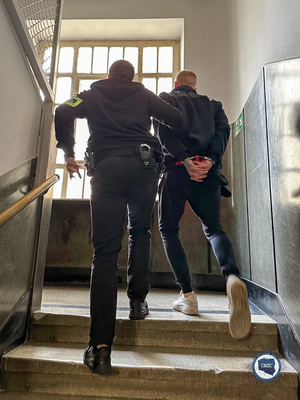 Policjant prowadzi schodami do góry zatrzymanego w kajdankach.
