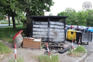 Spalona wiata śmietnikowa i nadpalone kontenery na śmieci.