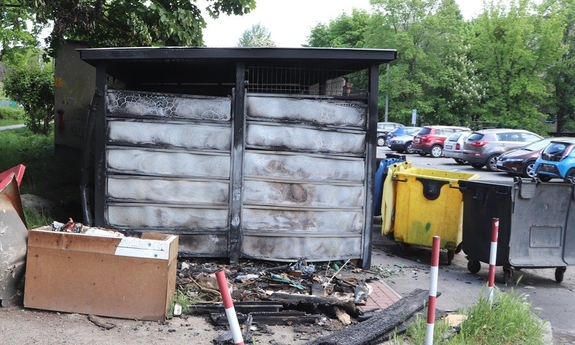 Spalona wiata śmietnikowa i nadpalone kontenery na śmieci.