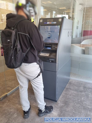 Mężczyzna w kasku stoi przy bankomacie.