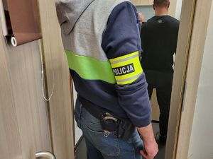 funkcjonariusz policji po cywilnemu z opaską Policja na ręku stojący w drzwiach pomieszczenia