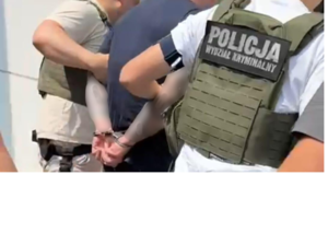 Dwaj policjanci w kamizelkach z napisem Policja na plecach prowadzą zatrzymanego.
