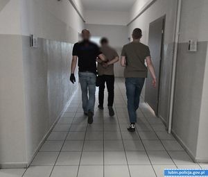 Dwaj policjanci w cywilnych ubraniach prowadzą skutego w kajdanki mężczyznę korytarzami komendy.