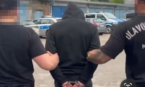 Dwóch mężczyzn prowadzi trzeciego mężczyznę, który ma założone kajdanki na ręce trzymane z tyłu.