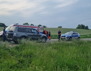 Policjanci prowadzą uratowanego mężczyznę do karetki. Z boku widoczny jest policyjny radiowóz i cywilne auto.