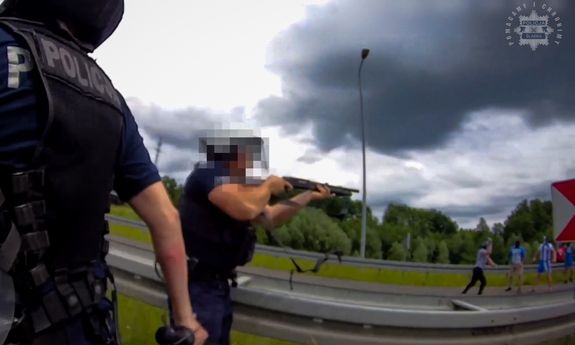Policjant trzyma w rękach broń. W oddali widać grupę kiboli.