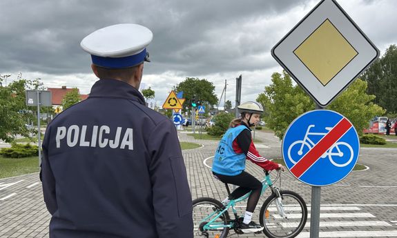 plecy policjanta, przed nim jeździ dziecko na rowerze.