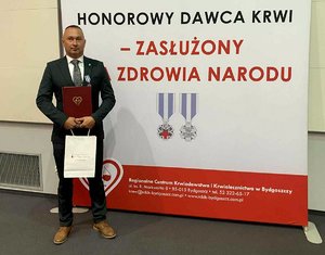 Odznaczony policjant w garniturze pozuje do zdjęcia na tle ścianki z napisem: Honorowy Dawca Krwi – Zasłużony dla Zdrowia Narodu.