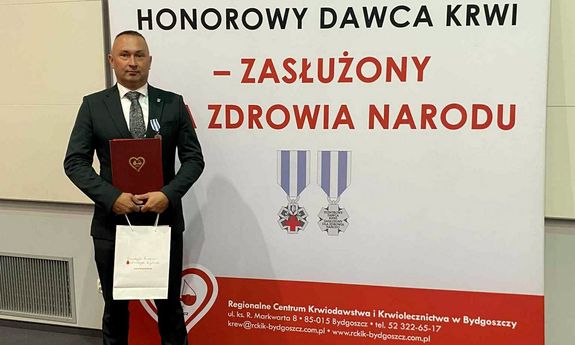 Odznaczony policjant w garniturze pozuje do zdjęcia na tle ścianki z napisem: Honorowy Dawca Krwi – Zasłużony dla Zdrowia Narodu.