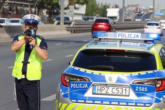 Umundurowany policjant stojący przy oznakowanym radiowozie w trakcie dokonywania pomiaru prędkości laserowym miernikiem prędkości