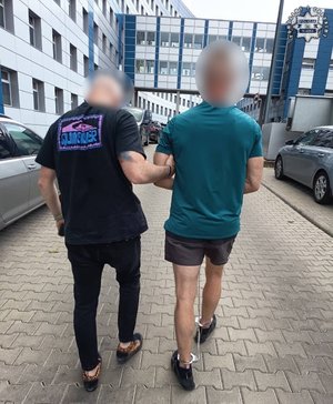 Na zdjęciu policjant w stroju cywilnym prowadzący zatrzymanego mężczyznę, który ma założone kajdanki zespolone.
