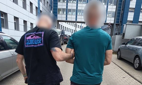 Na zdjęciu policjant w stroju cywilnym prowadzący zatrzymanego mężczyznę, który ma założone kajdanki zespolone.