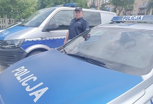 Umundurowany policjant stoi na tle oznakowanych radiowozów.