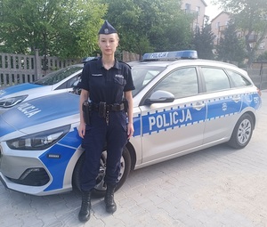 Umundurowana policjantka stoi na tle oznakowanego radiowozu.